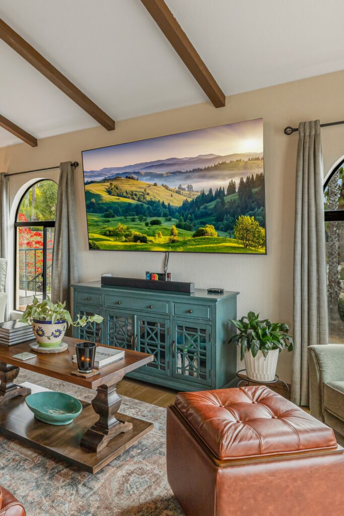Living room with vaulted wood beams, arched windows, wall-mounted TV, teal media console and leather ottoman.