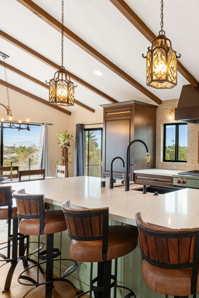 Green kitchen island with white counters, four barstools, black-and-brass faucets, pendants and vaulted wood beams.