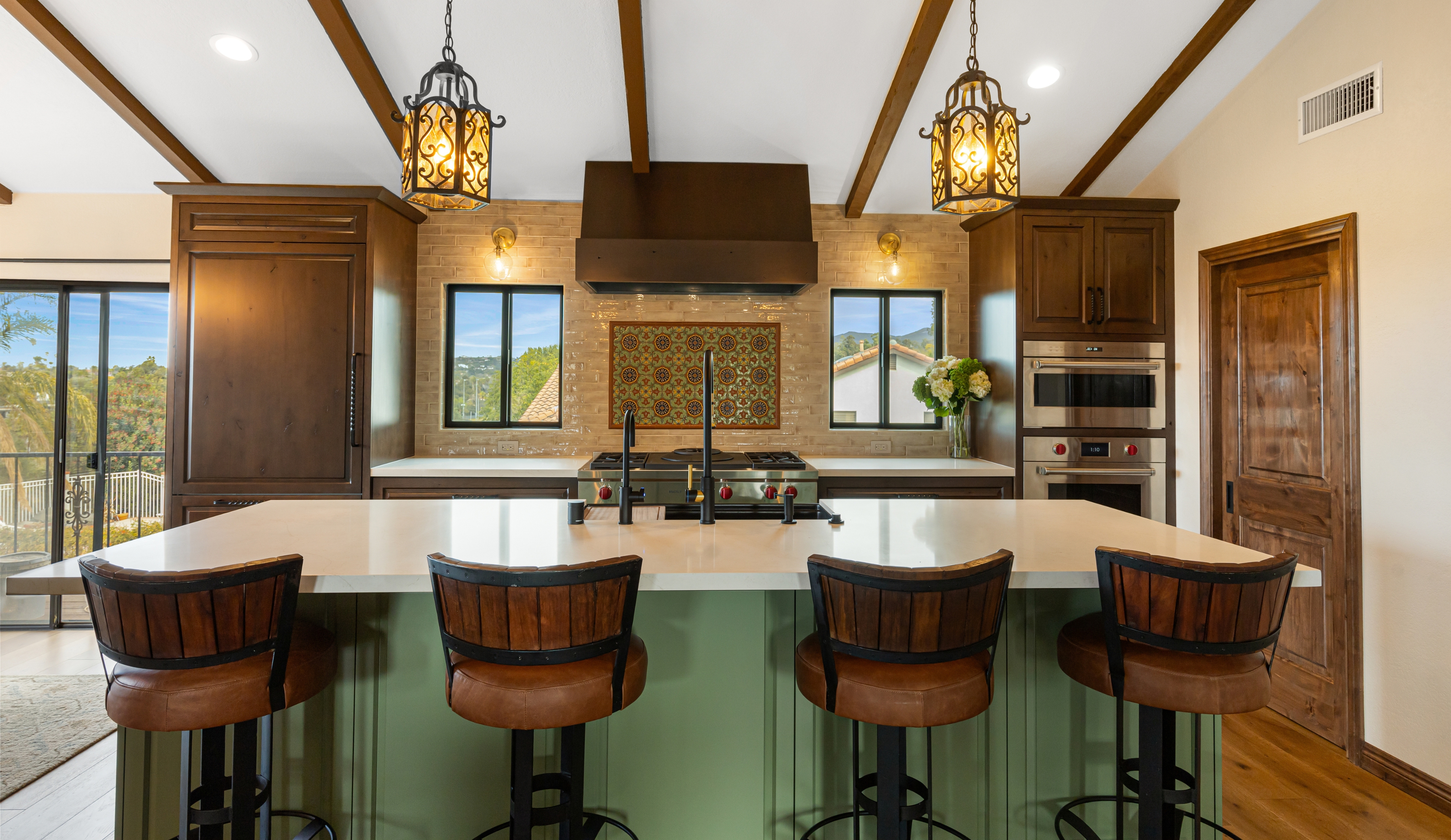 Open Vista kitchen with green island seating, dark wood cabinets, exposed beams, range wall and Spanish tile mural.