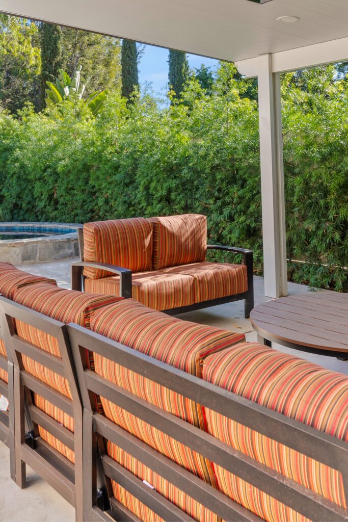 Covered outdoor lounge with striped seating, round table, concrete patio and dense garden screening.