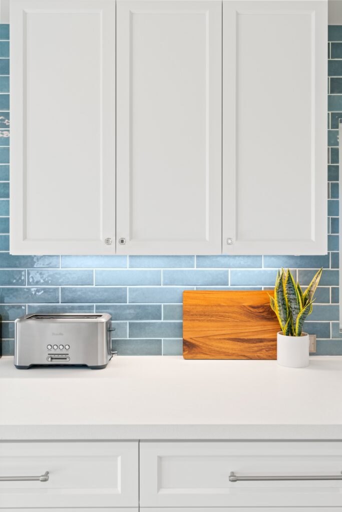 White upper and lower cabinets with blue subway tile, white countertop and stainless toaster.