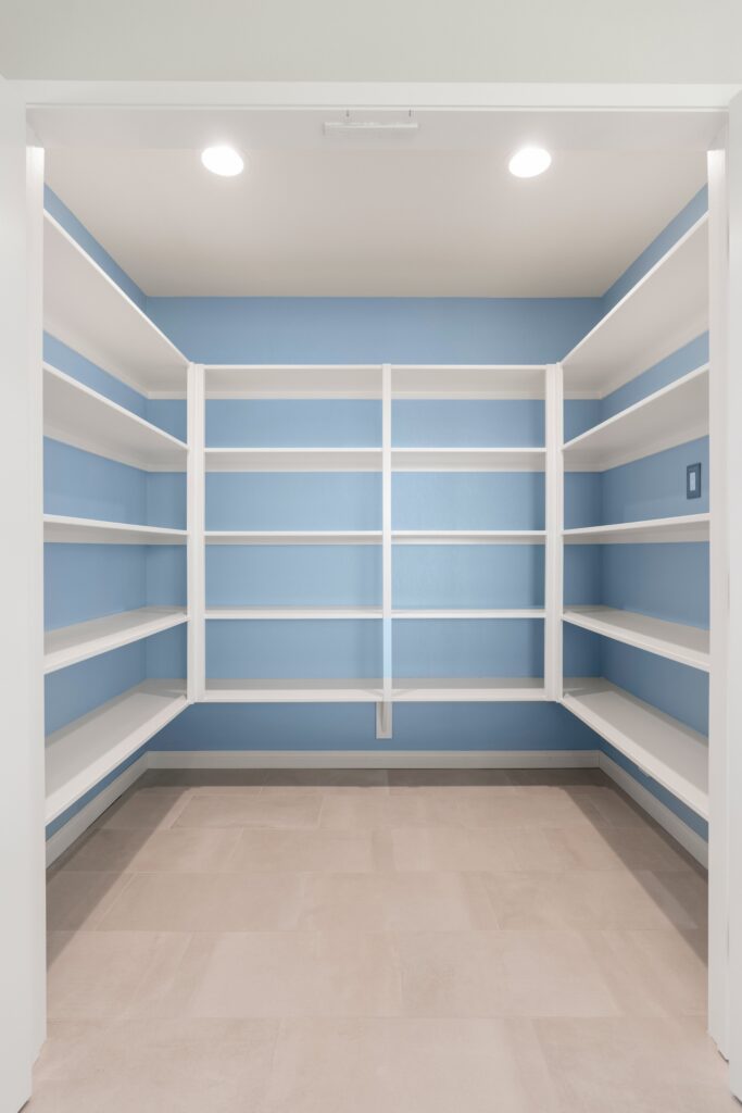 Walk-in pantry with blue walls, white wraparound shelving, recessed lights and light tile flooring.