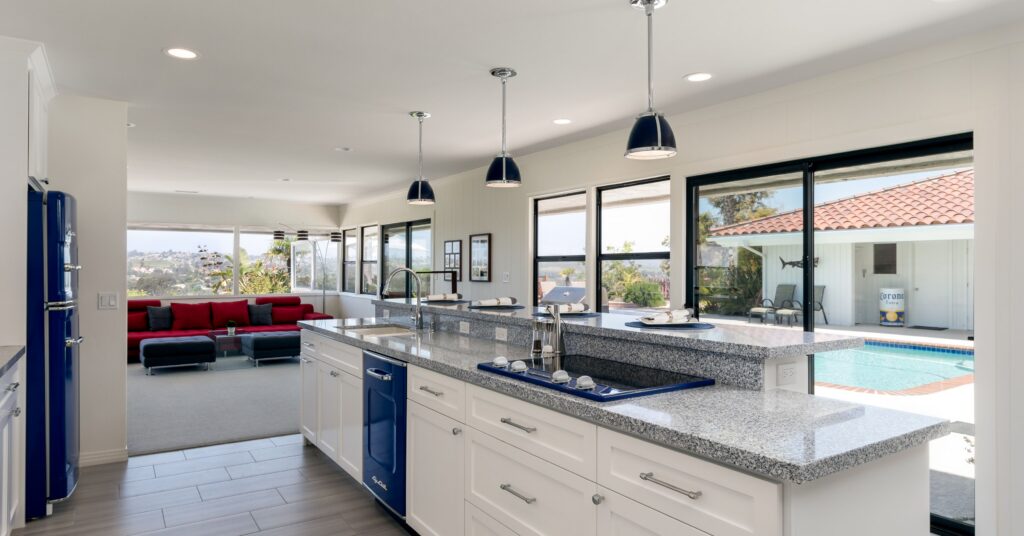 Open-concept kitchen remodeled through a design-build process featuring white shaker cabinets, granite countertops, blue retro appliances, a large island, and expansive windows overlooking the backyard pool.