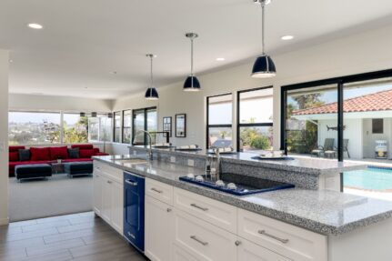 Open-concept kitchen remodeled through a design-build process featuring white shaker cabinets, granite countertops, blue retro appliances, a large island, and expansive windows overlooking the backyard pool.