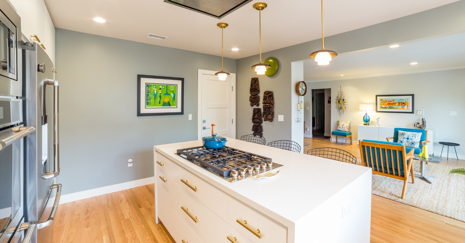 Open kitchen with gray walls, a white cooktop island, brass pendant lights, stainless steel appliances, and an adjoining living room.