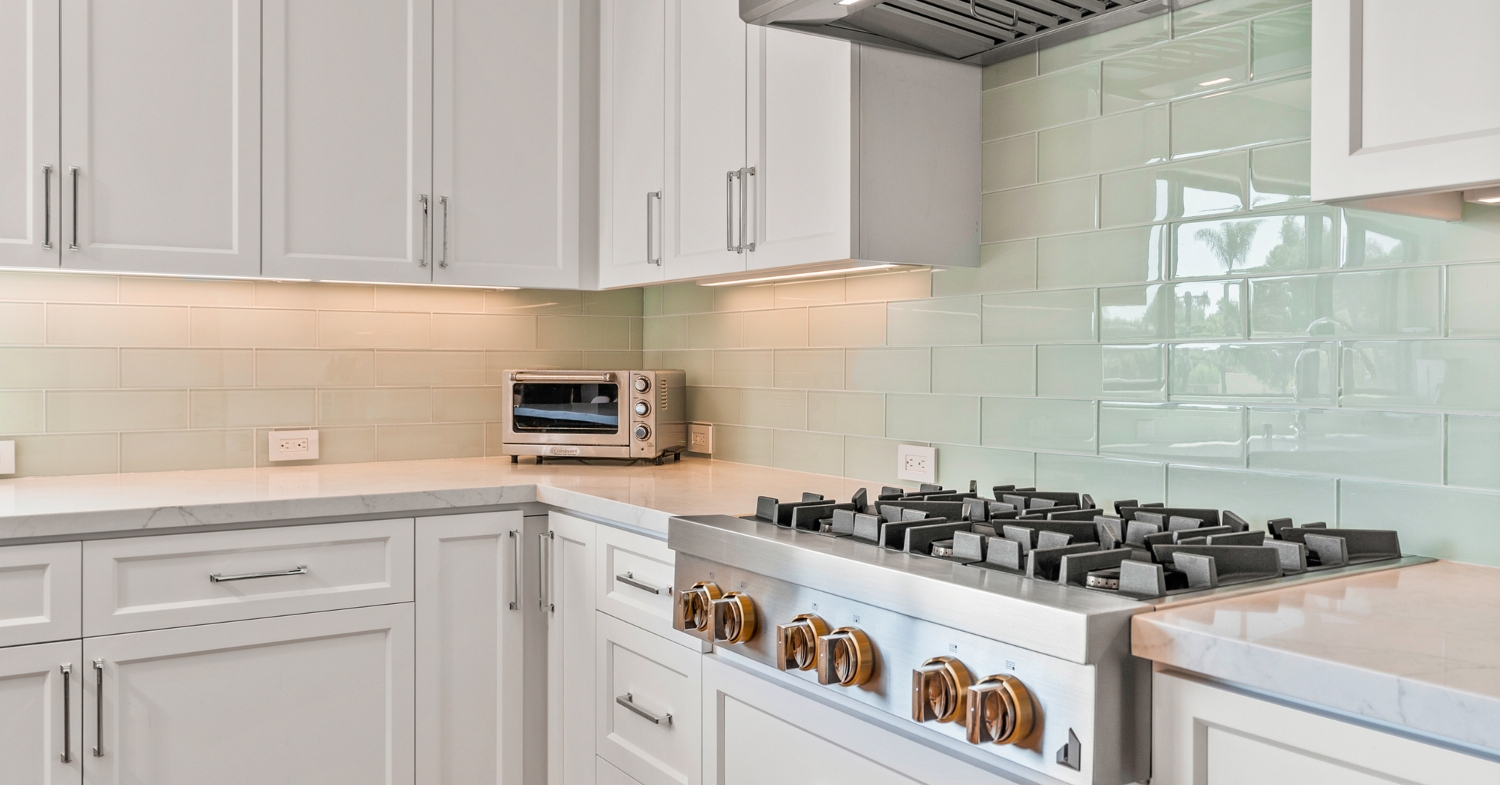 White shaker kitchen with a stainless professional gas cooktop, marble-look quartz counters and a glossy sea-glass subway tile backsplash.