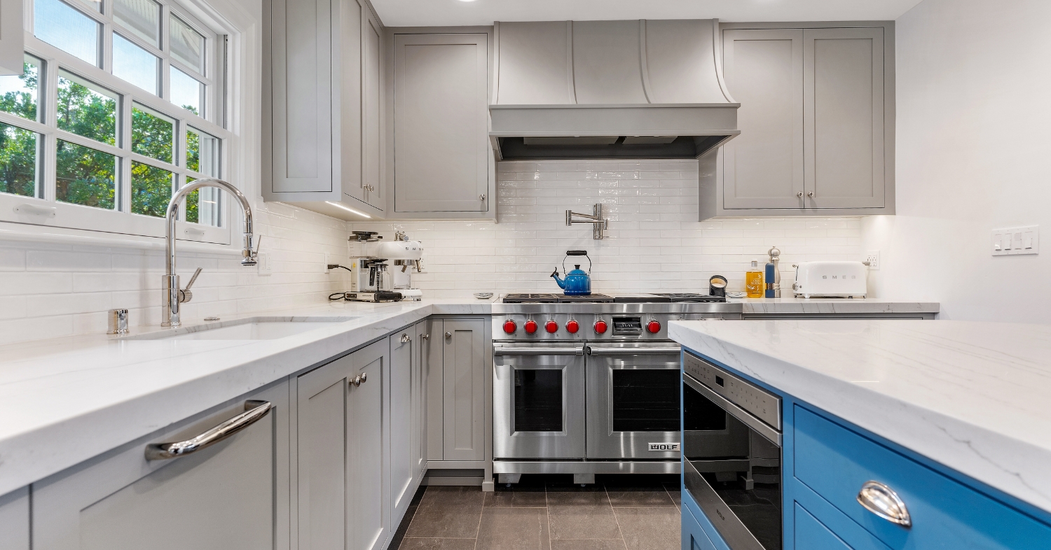 Compact gray kitchen with white countertops, a blue island, a professional range, and a sink positioned beneath a large window.