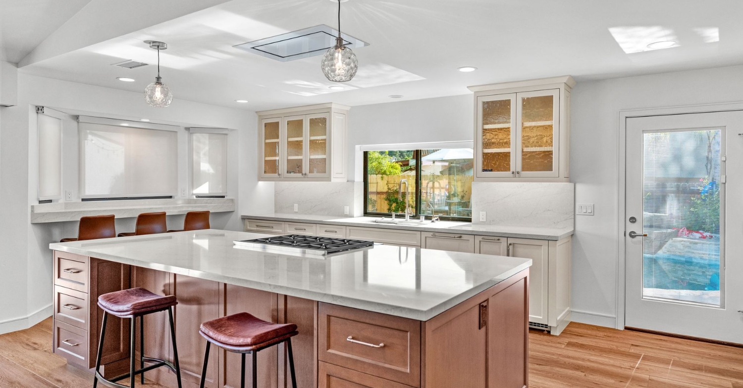 Remodeled kitchen with a brown wood island, white perimeter cabinets, quartz countertops, and a cooktop facing a backyard window.