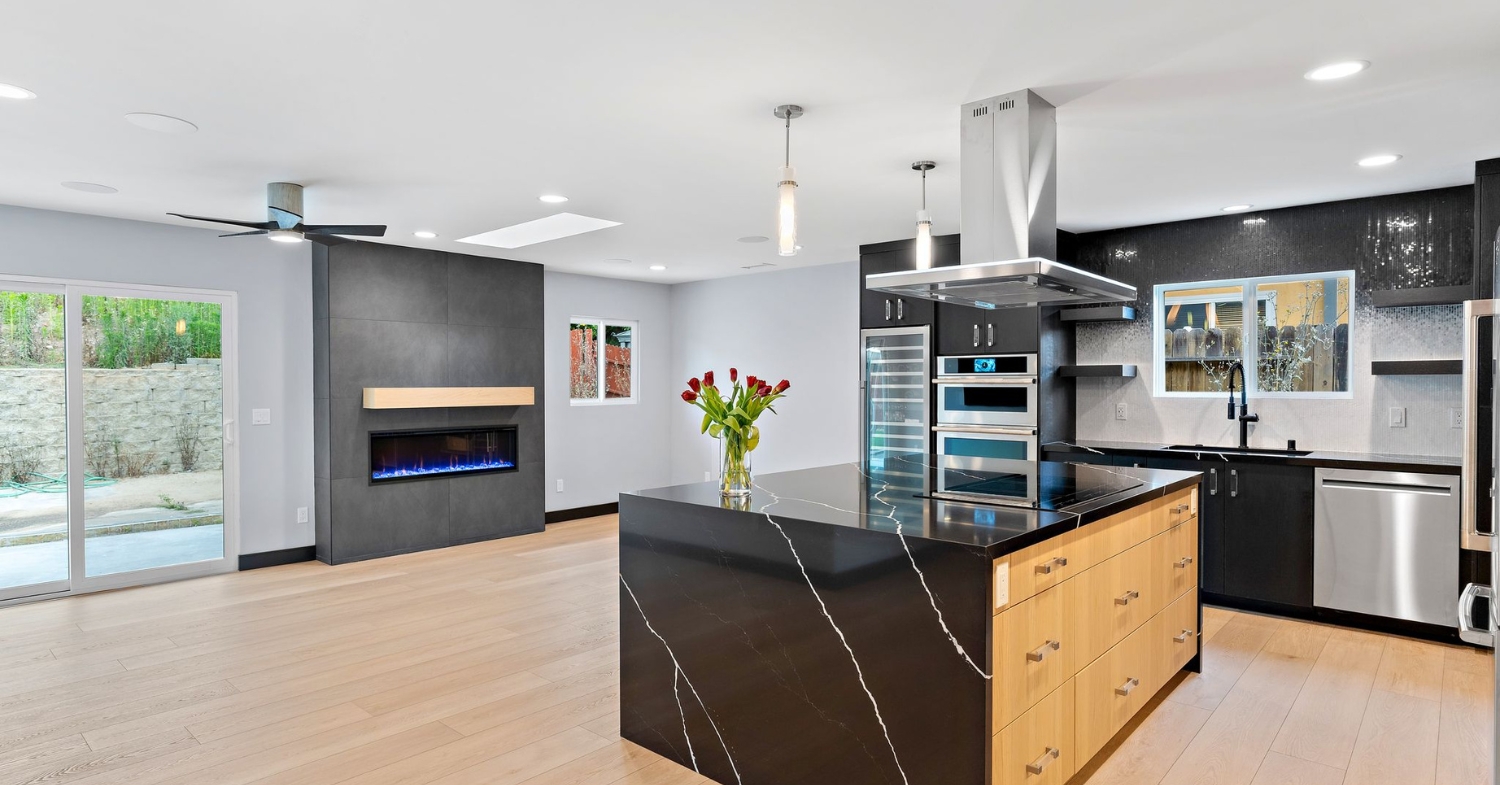 Open-concept remodeled kitchen and living space with a waterfall quartz island, black cabinetry, white oak flooring and a linear fireplace.