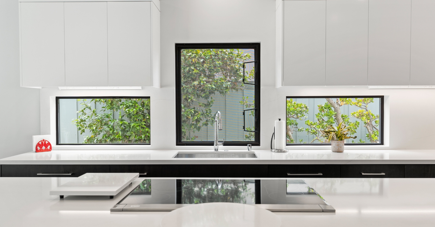 Minimalist white kitchen with handleless slab cabinets, waterfall quartz counters, black-framed windows and an integrated cooktop.