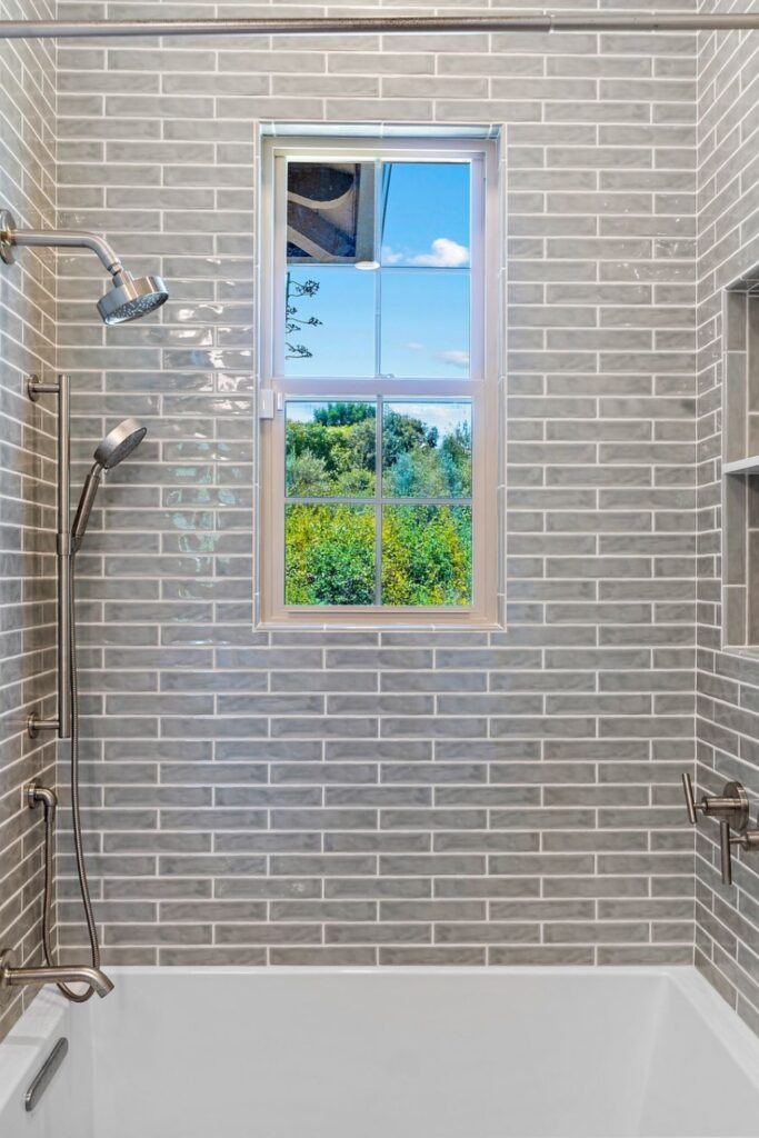 Gray tiled bathtub and shower with a window, handheld shower fixture, and brushed-nickel plumbing hardware.
