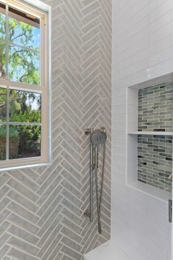 Gray herringbone tile shower wall with a window, handheld shower, and built-in mosaic tile niches.
