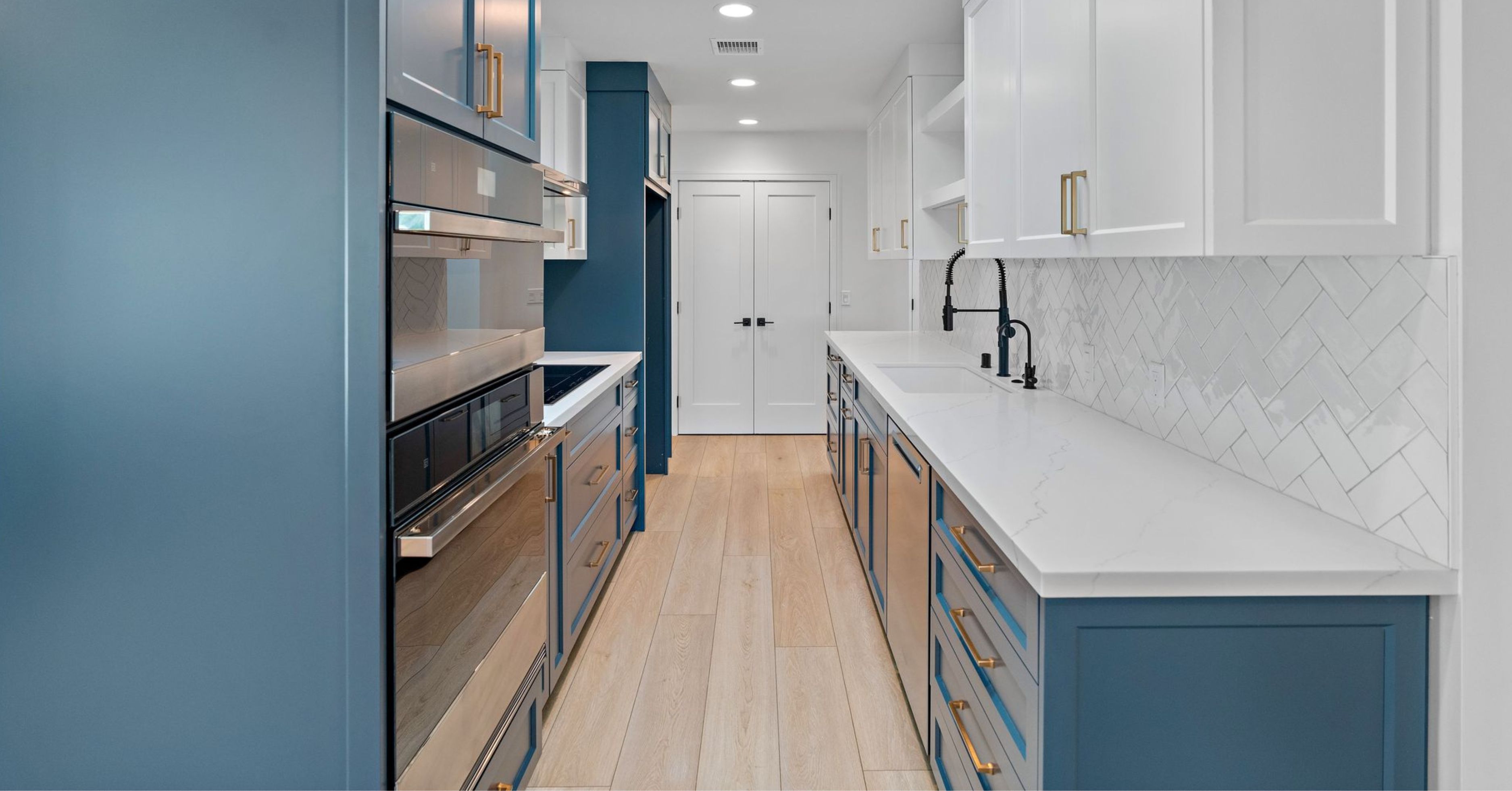 Modern galley kitchen remodel with blue and white custom cabinetry, quartz countertops, herringbone tile backsplash, and built-in stainless steel appliances.