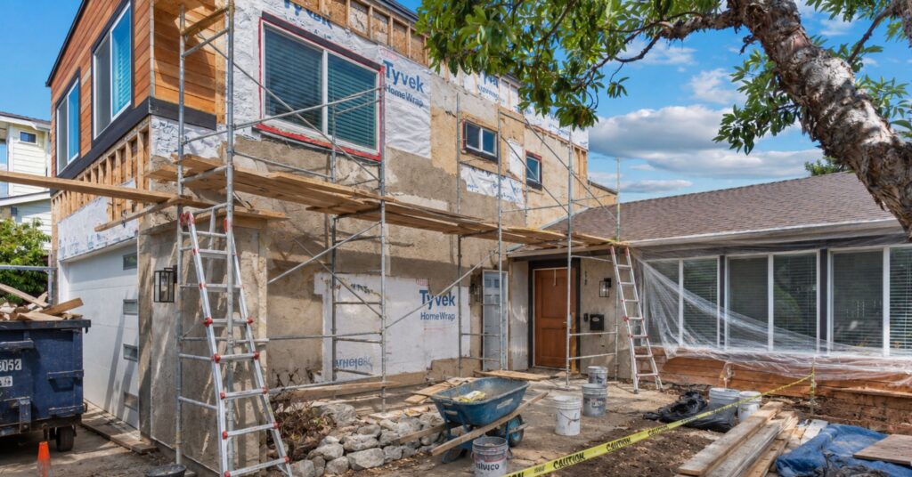 Two-story San Diego home undergoing an exterior remodel with scaffolding, ladders, house wrap, lumber, and construction materials.