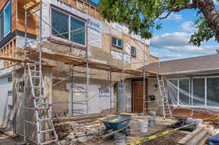 Two-story San Diego home undergoing an exterior remodel with scaffolding, ladders, house wrap, lumber, and construction materials.