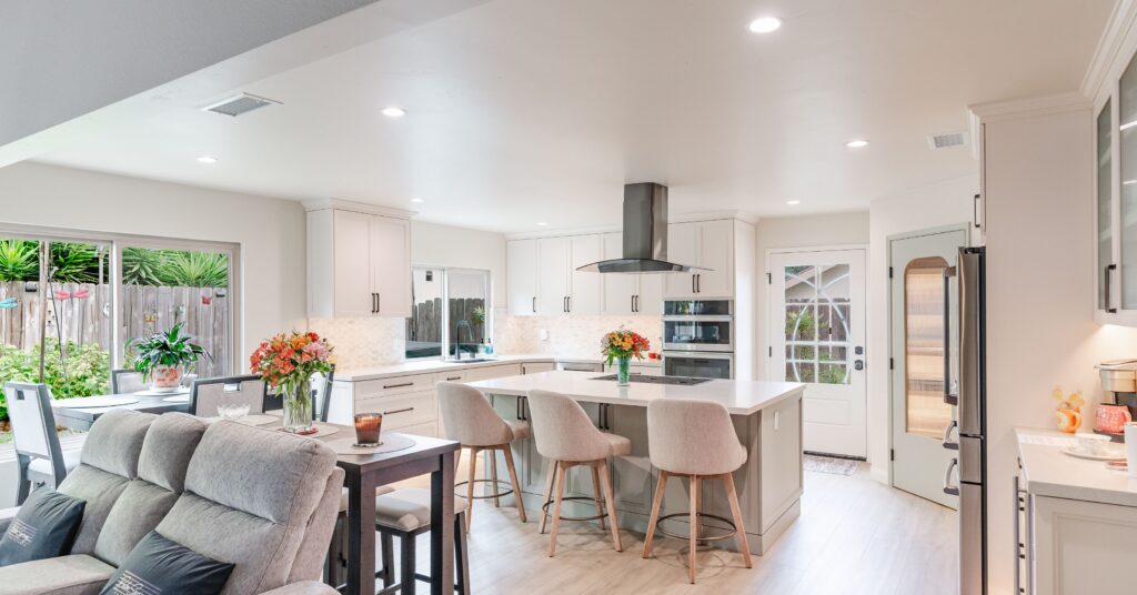 Open-concept kitchen and living area with a large island, four upholstered stools, white cabinetry, and sliding glass doors to the backyard.