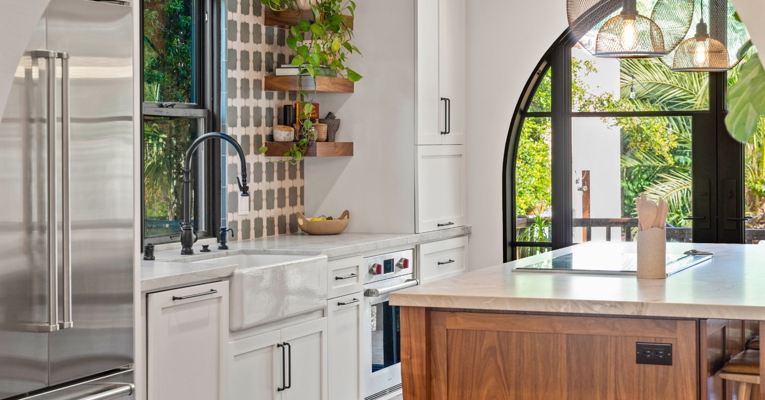 Transitional white kitchen with a farmhouse apron sink, patterned tile backsplash, walnut island, marble-look counters and an arched black-framed window.
