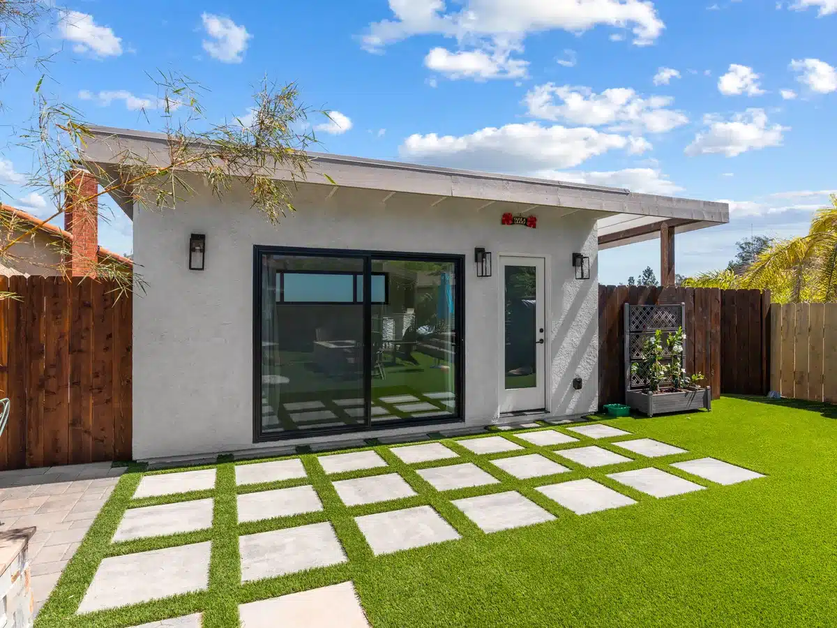 Detached backyard building with a dark sliding glass door, white exterior and a geometric paver path.