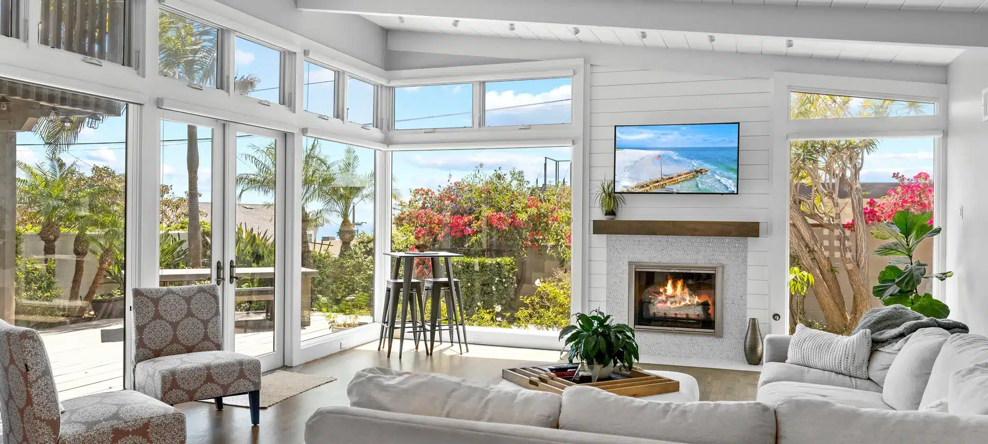 Sunlit living room with a fireplace, exposed ceiling beams and large windows facing a planted patio.