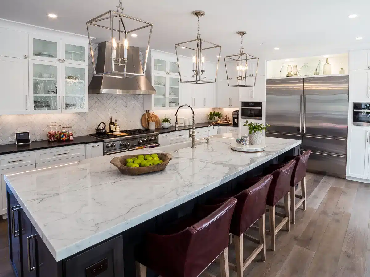 White kitchen with a broad stone island, counter seating and glass pendant lights.