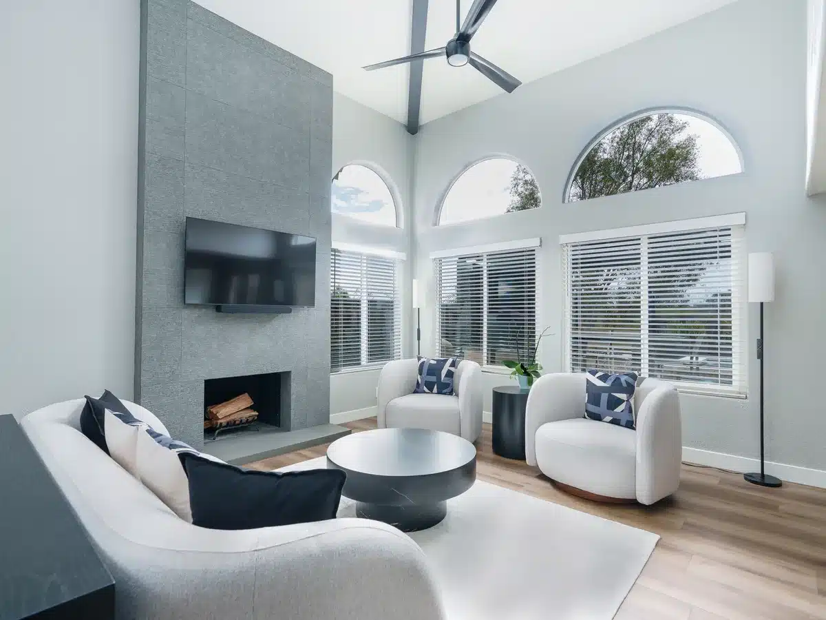 Living room with a vaulted ceiling, arched windows, light seating and a tall gray fireplace wall.
