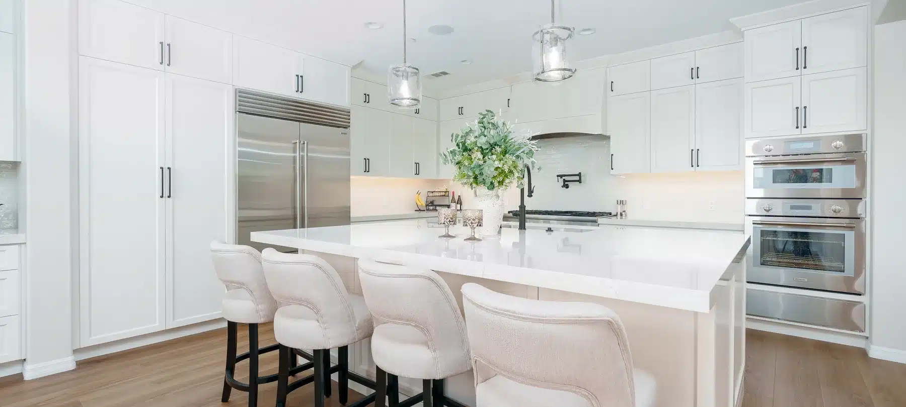 White kitchen with a wide island, upholstered stools, and pendant lights.