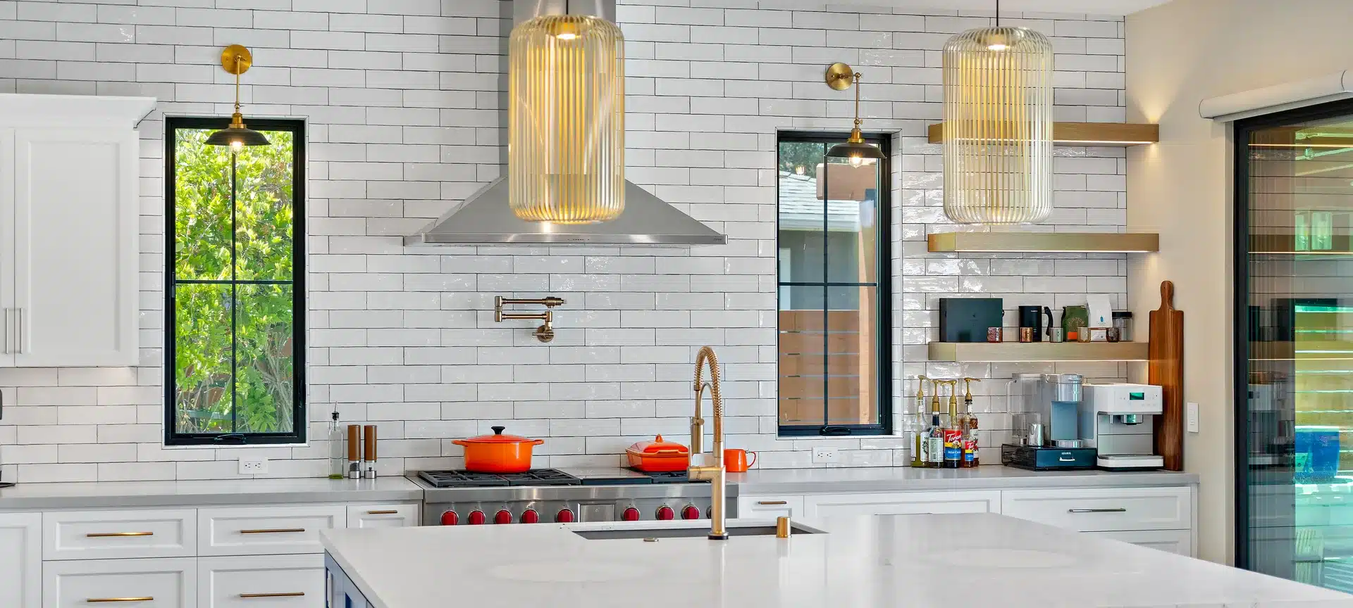 Kitchen with white tile walls, black-framed windows, ribbed brass pendant lights, and a pale island countertop.