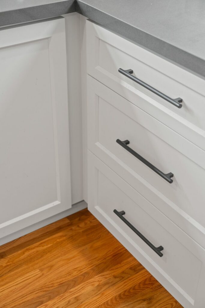 White kitchen drawers and corner cabinet with dark bar pulls, gray countertop and refinished hardwood flooring.