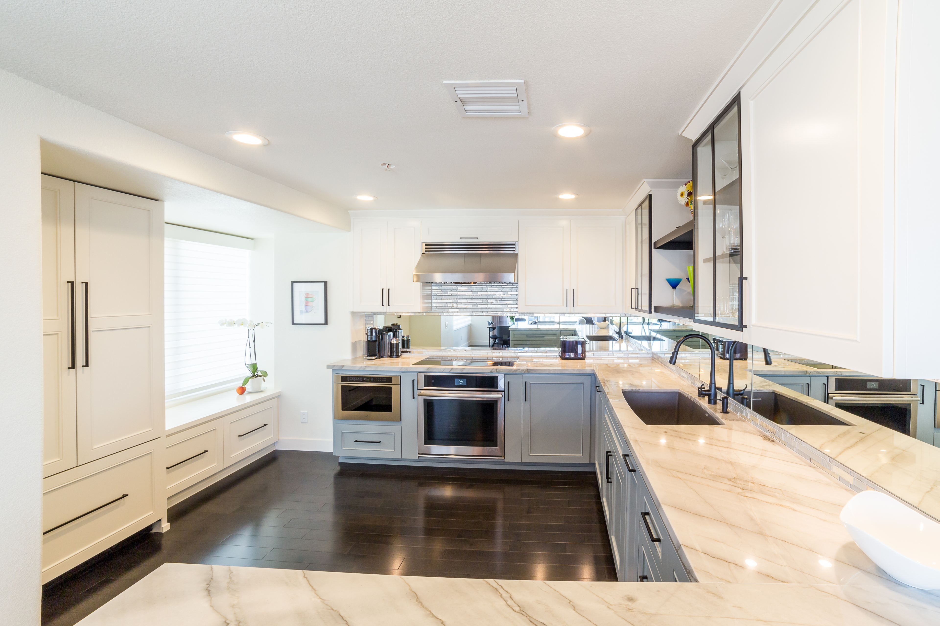 White and gray kitchen with stone counters, mirrored backsplash, window bench, glass cabinets and dark wood flooring.