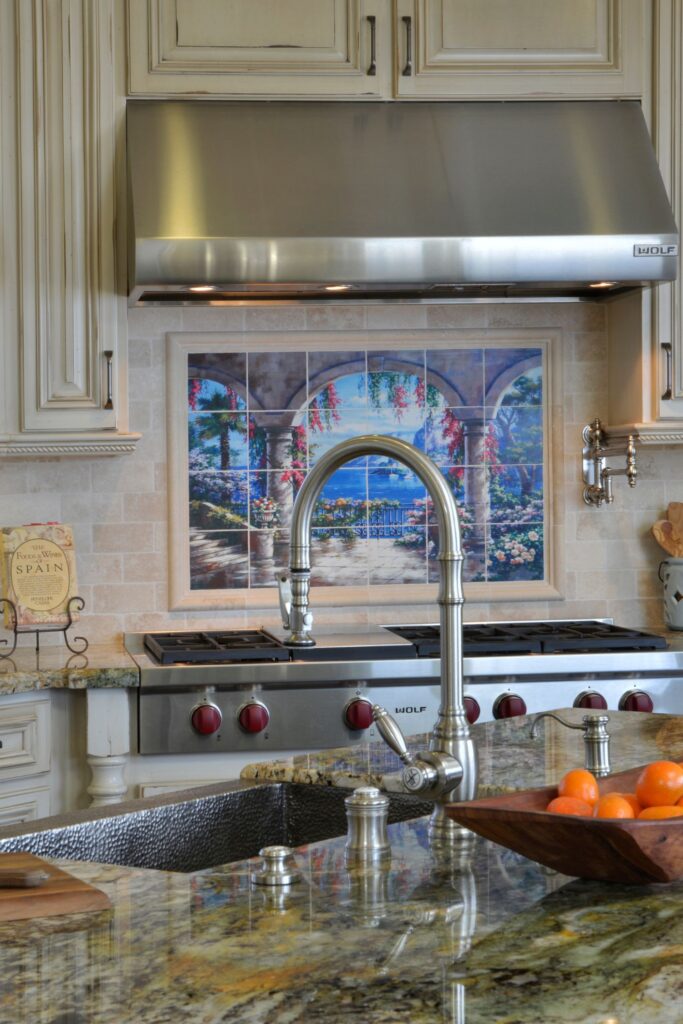 Kitchen range wall with stainless Wolf hood, scenic tile mural, cream cabinetry and granite island sink.