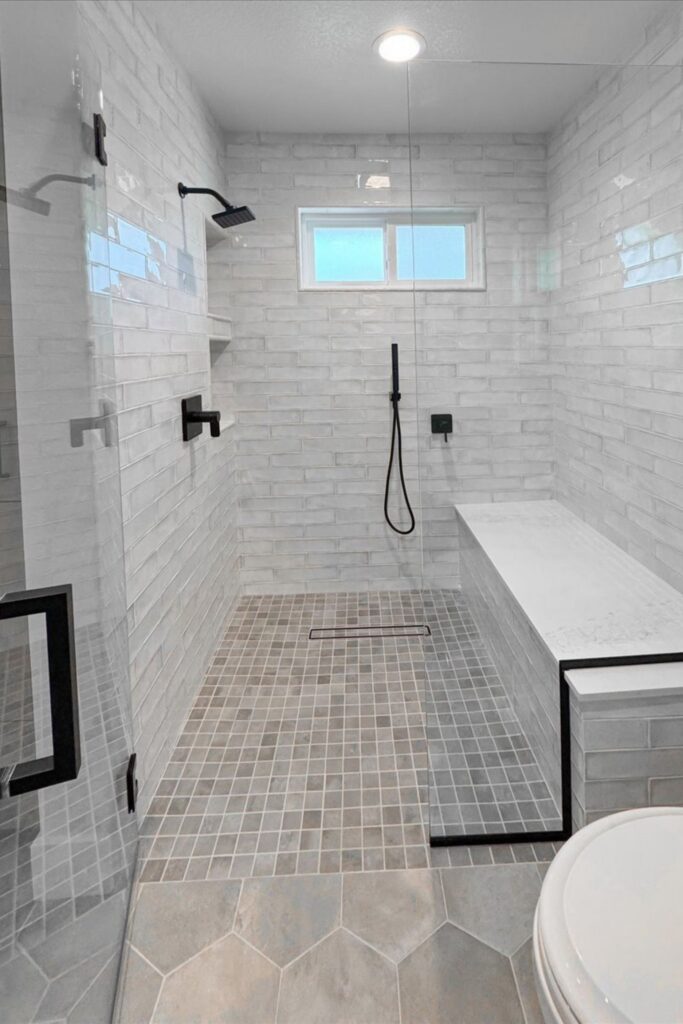 Walk-in shower with white subway tile, gray mosaic floor, built-in bench, black fixtures and frameless glass.
