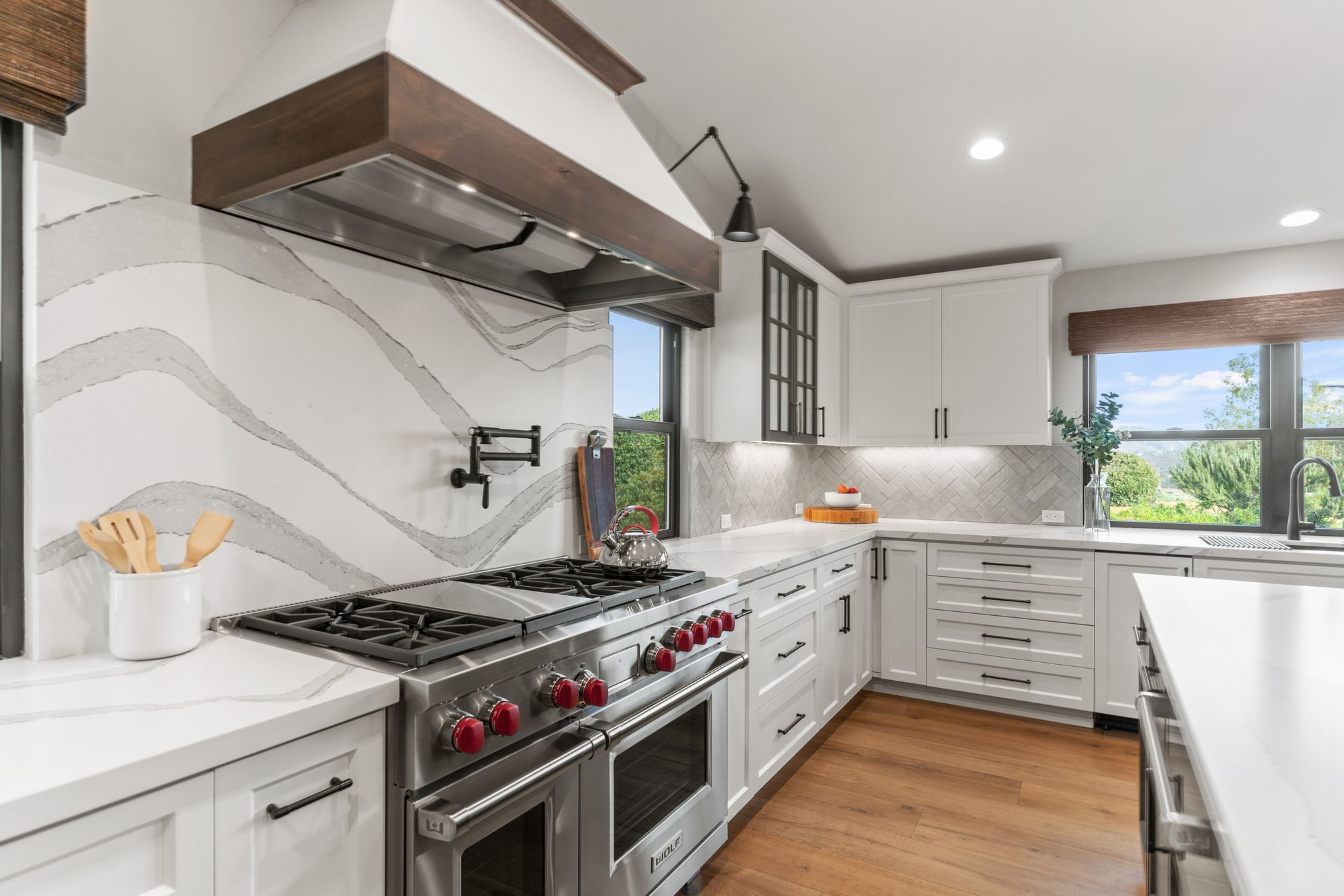 Encinitas remodeled kitchen with white cabinetry, Wolf range, quartz backsplash, wood-trimmed hood and black fixtures.