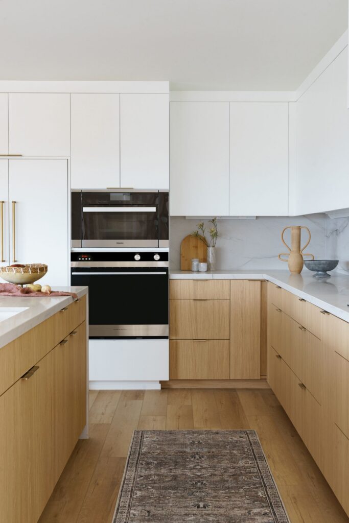 Kitchen with light wood lower cabinetry, white uppers, stacked ovens and marble-look counters and backsplash