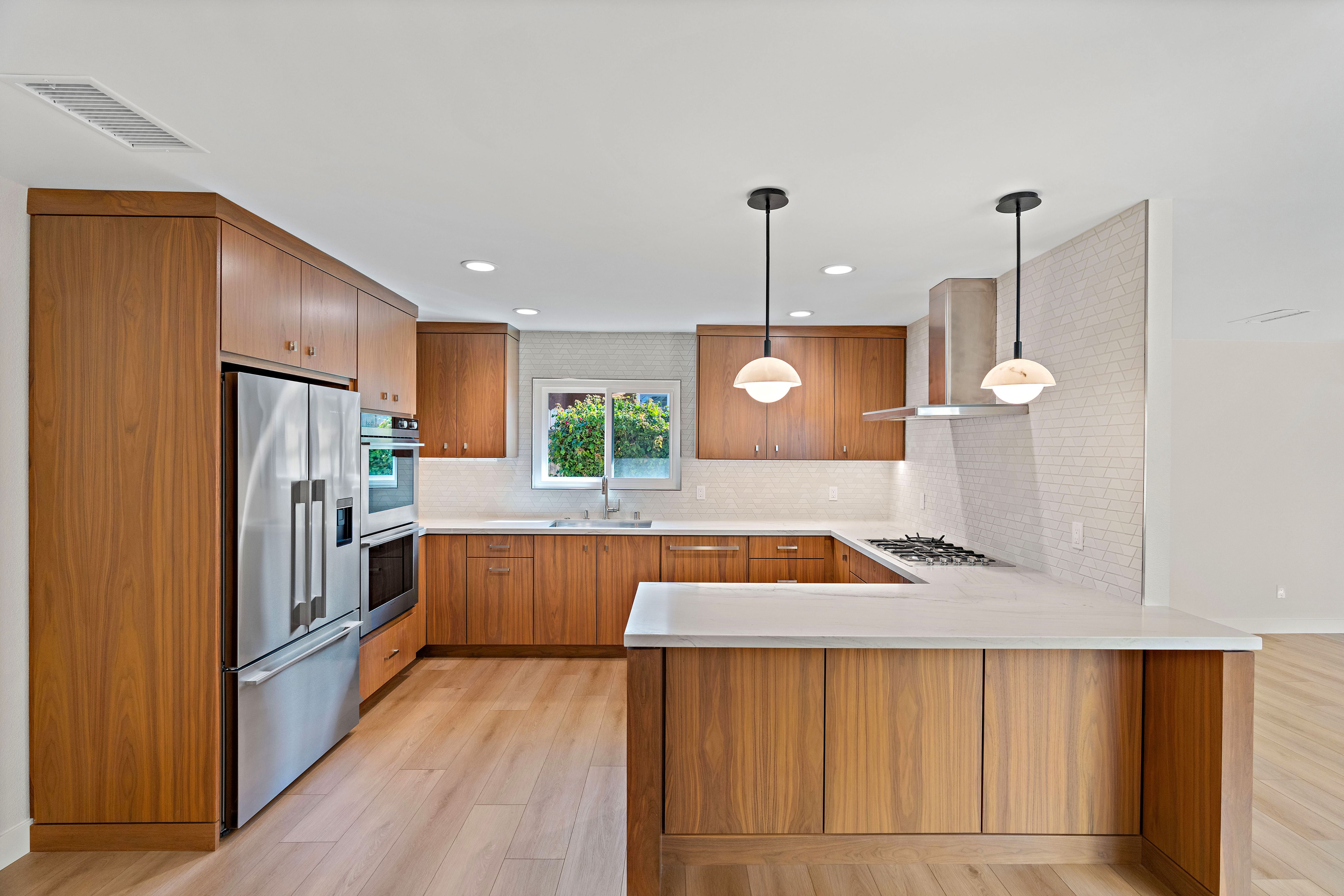 Open kitchen with walnut cabinetry, white counters, peninsula seating, geometric tile backsplash and pendant lights.