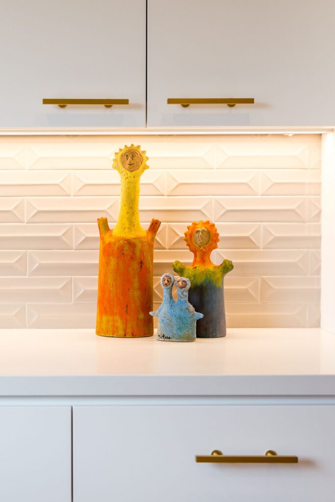 White kitchen cabinetry with brass pulls, dimensional white tile backsplash and colorful ceramic sculptures