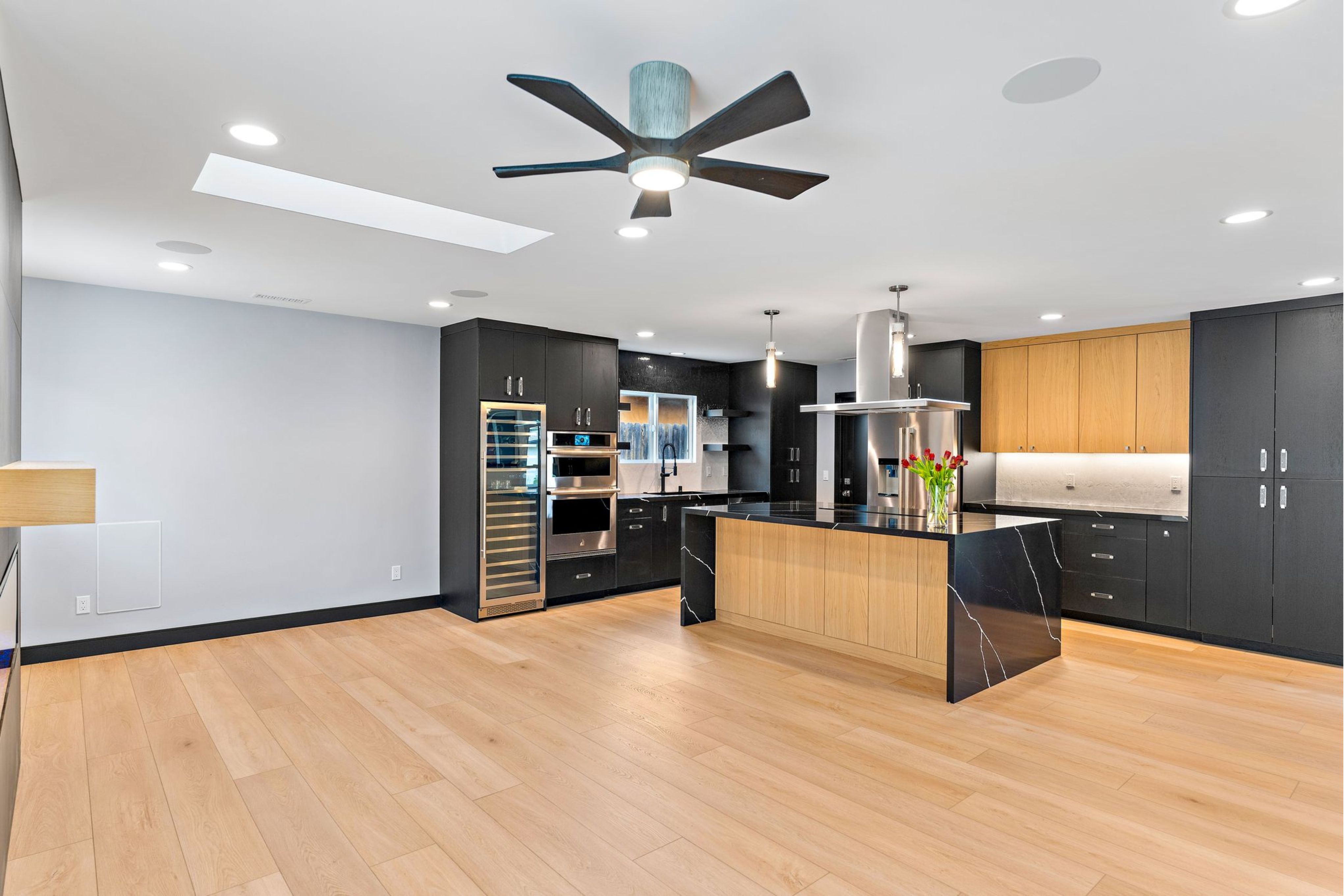 Open kitchen and living area with black cabinetry, wood accents, waterfall island, skylight and light wood flooring