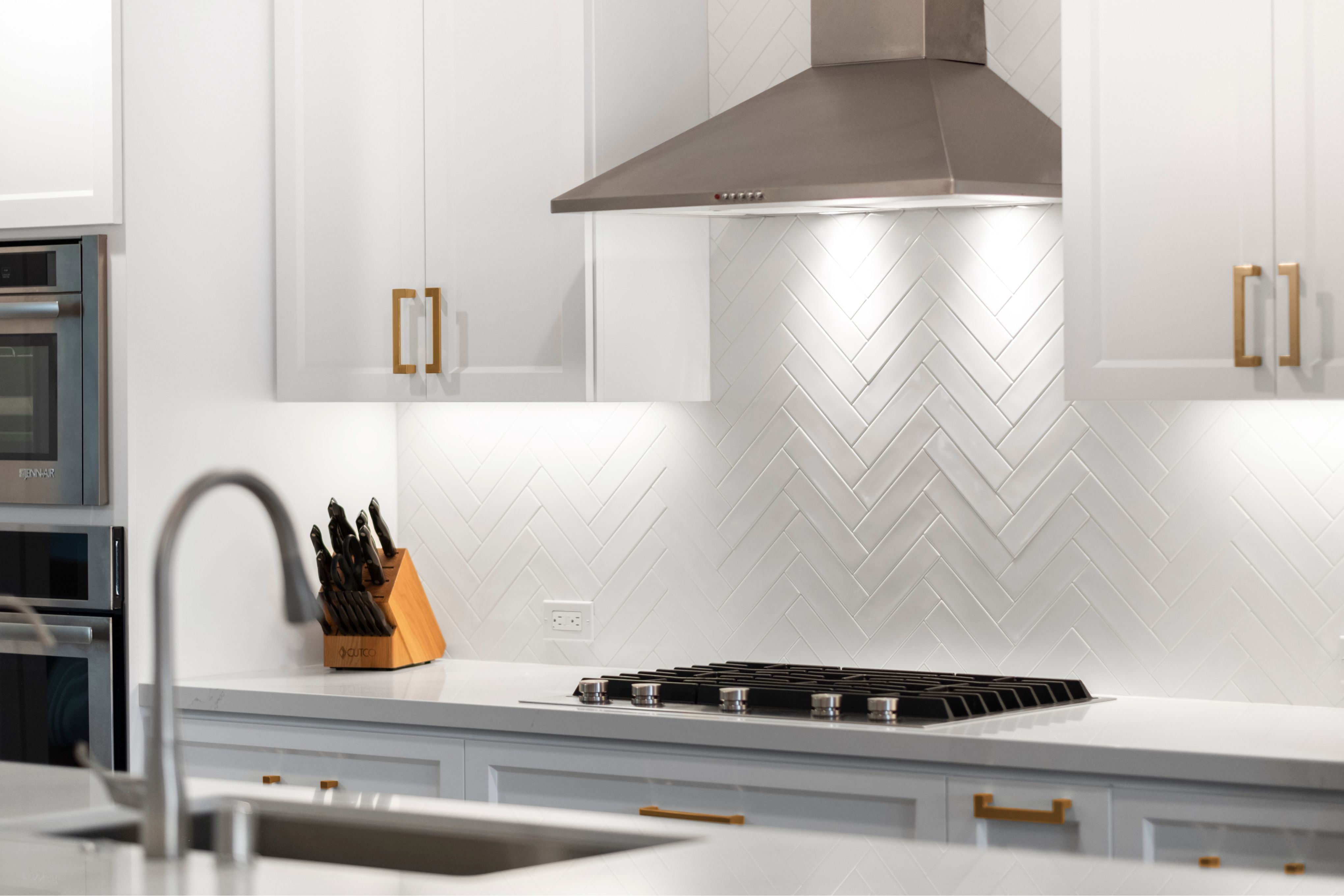 White kitchen with herringbone tile backsplash, stainless hood, cooktop, quartz counters, and brass hardware.