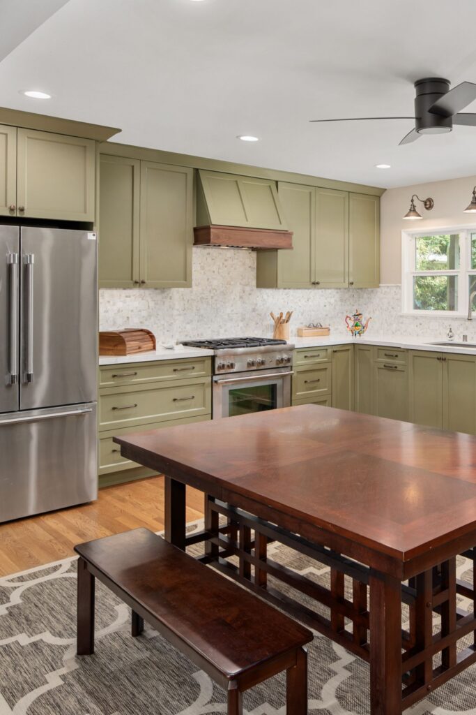 Sage green kitchen with stainless appliances, mosaic backsplash, wood dining table, and recessed lighting.