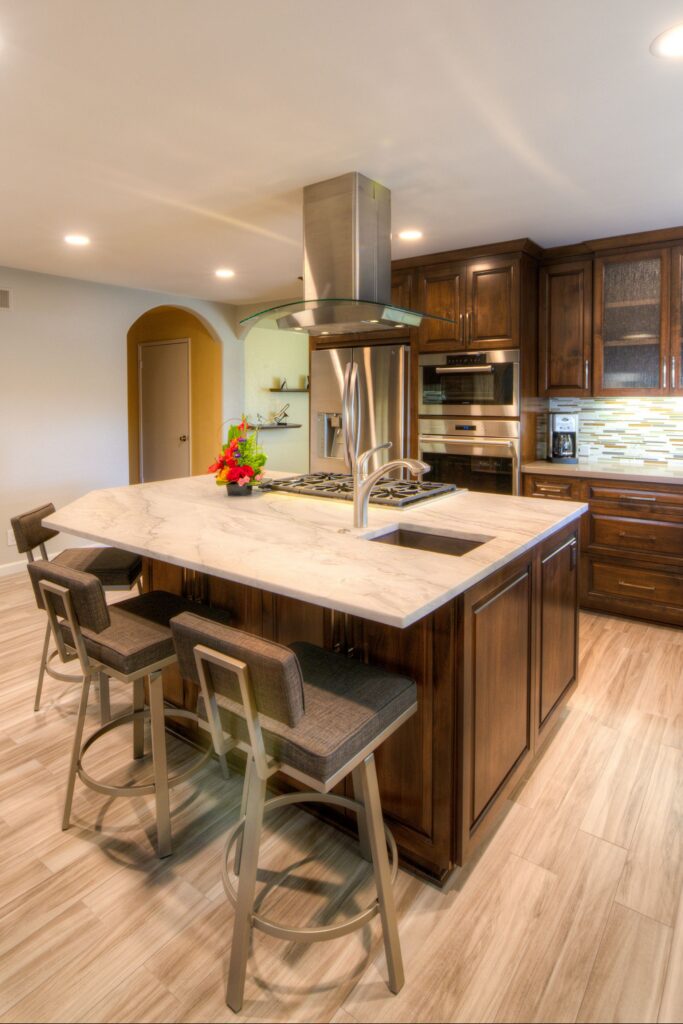 Dark wood kitchen island with cooktop, prep sink, stone-look countertop, seating and curved glass range hood.