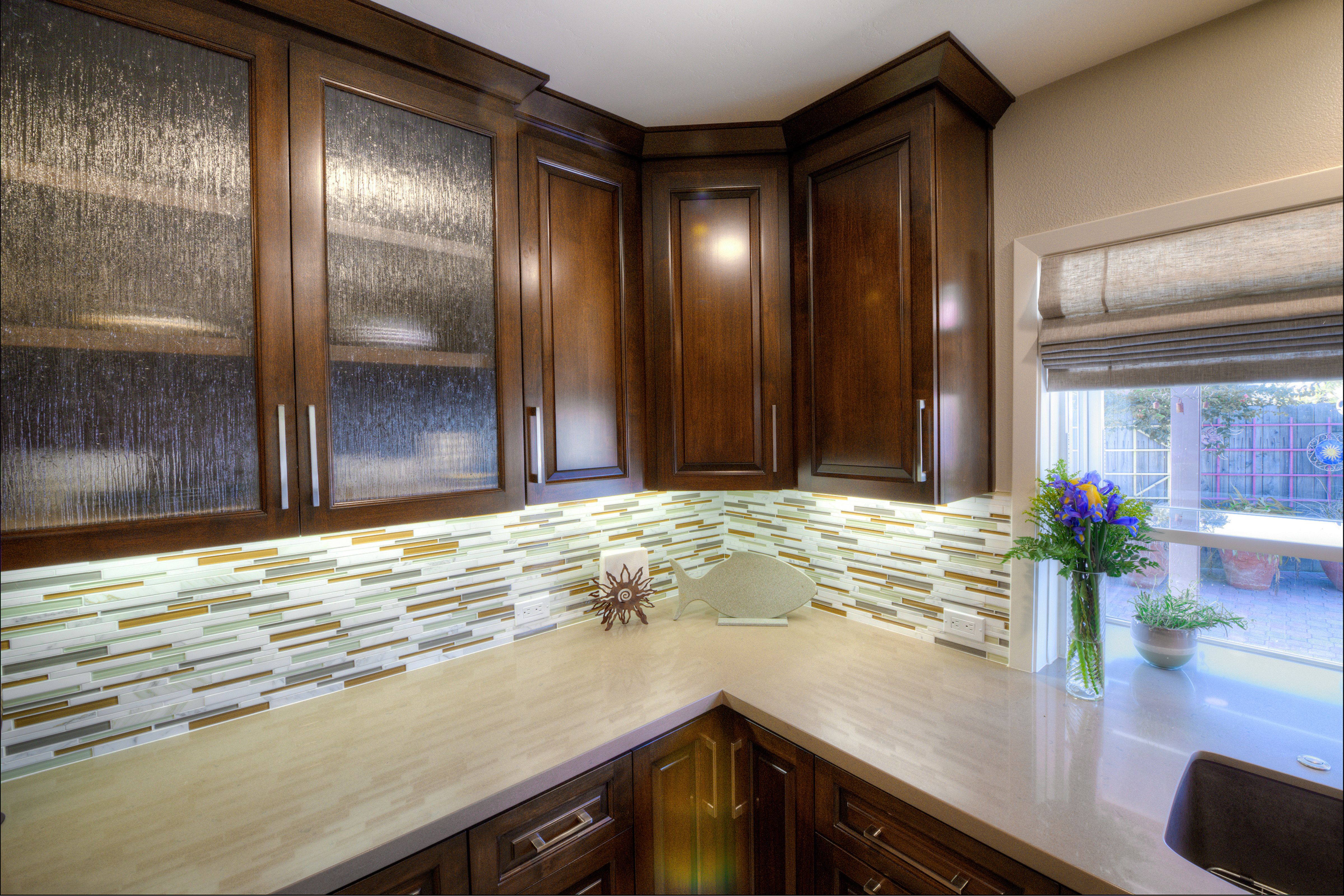 Dark wood kitchen cabinetry with textured glass doors, mosaic backsplash, light counters and under-cabinet lighting.