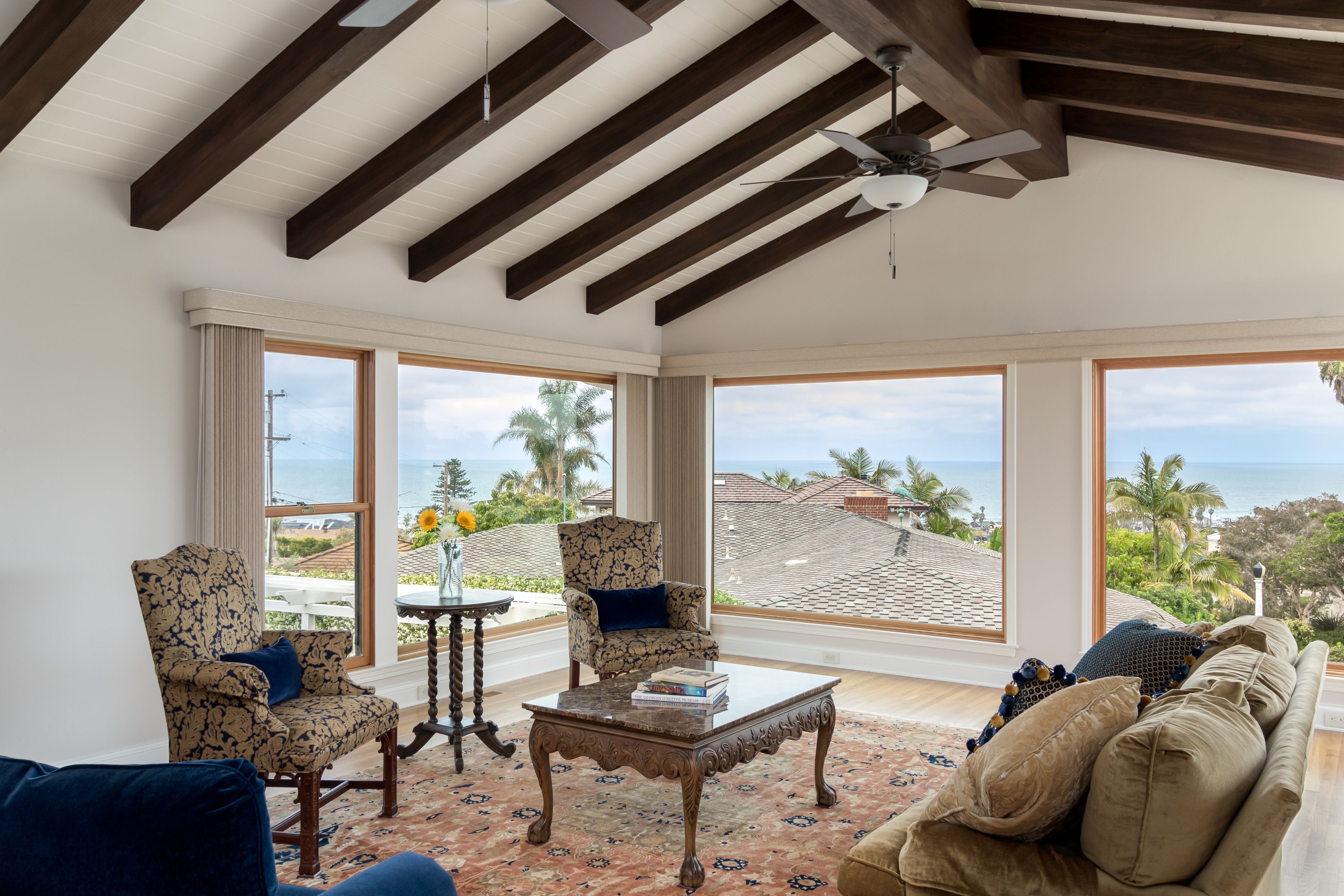 Vaulted living room with dark wood beams, white plank ceiling, wood-trimmed windows and ocean views.