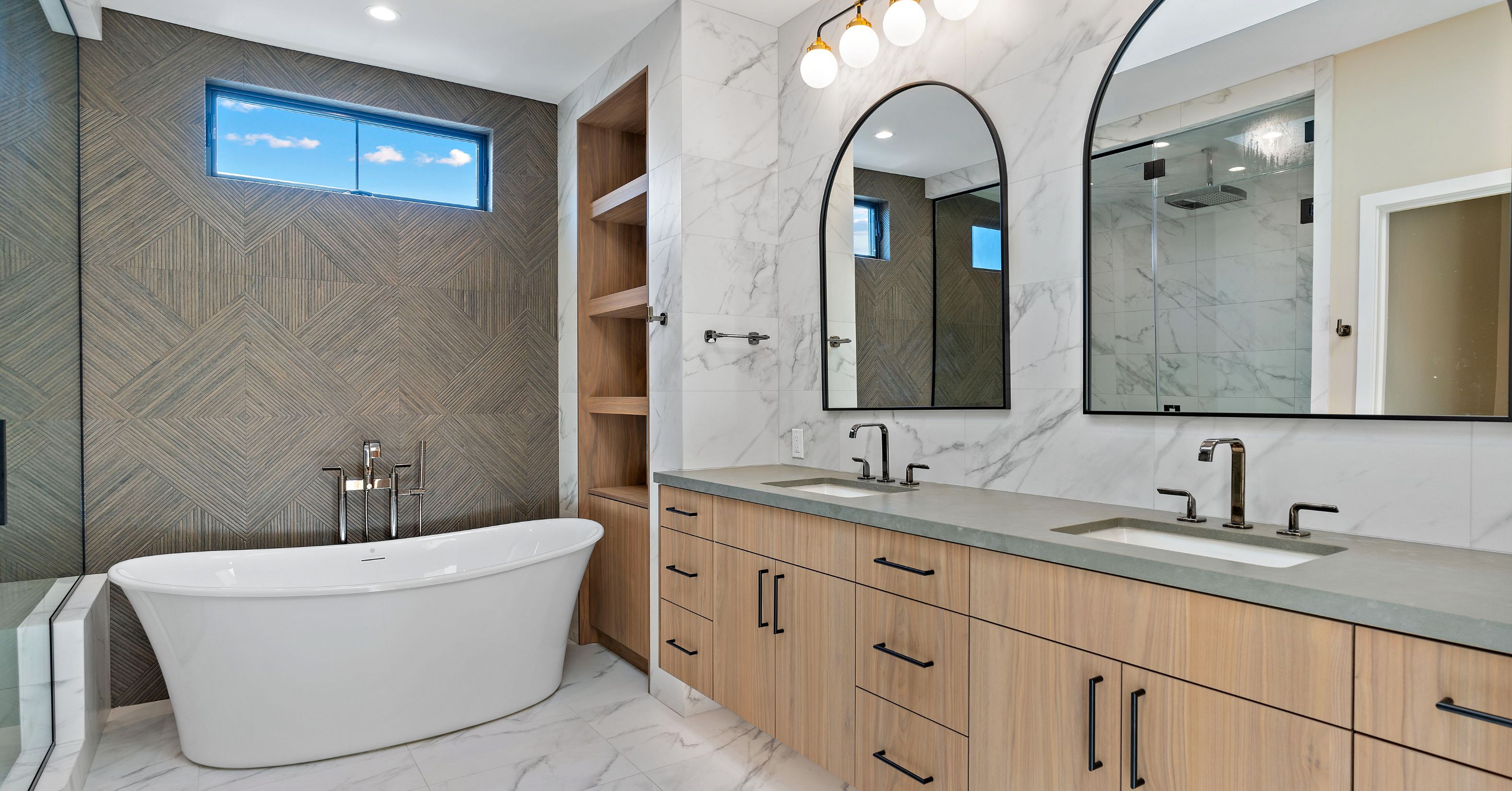 Primary bathroom with freestanding tub, double wood vanity, arched mirrors, marble-look tile, and glass shower.