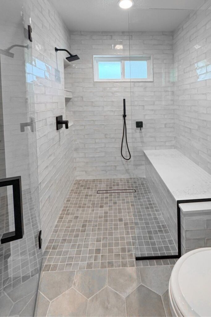 Walk-in shower with white wall tile, mosaic floor, built-in bench, black fixtures and frameless glass enclosure.
