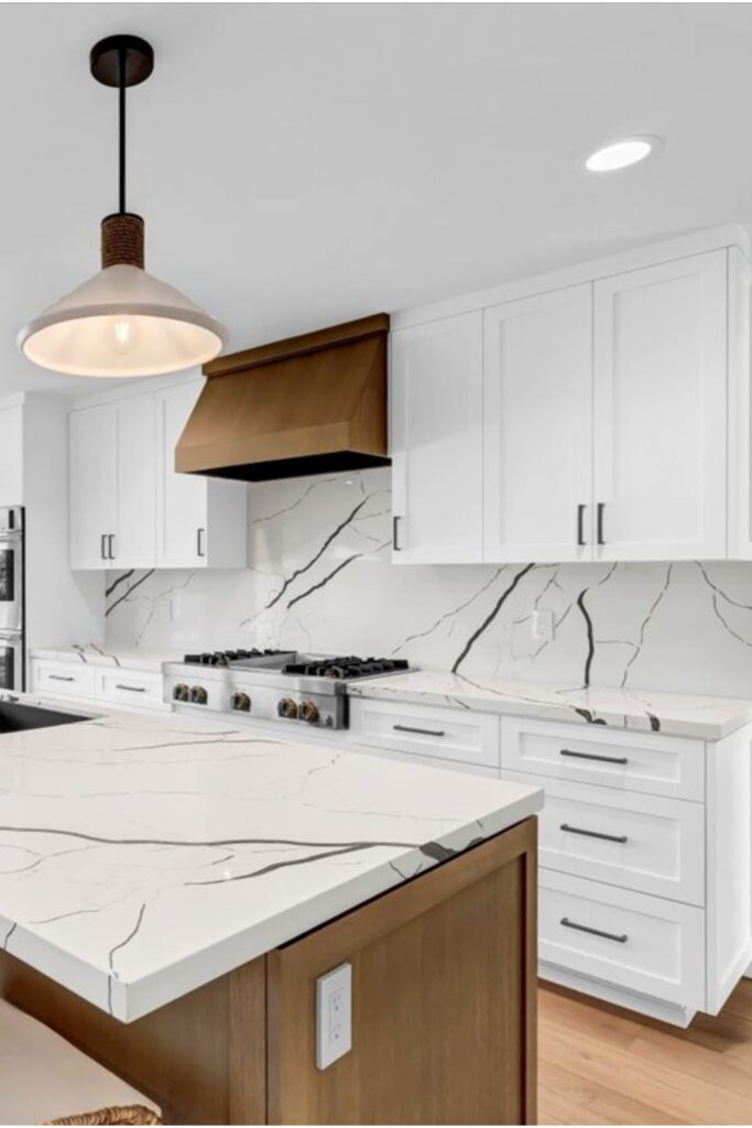 Remodeled kitchen with white cabinetry, wood island, veined slab countertops, brass-toned hood and pendant lighting.