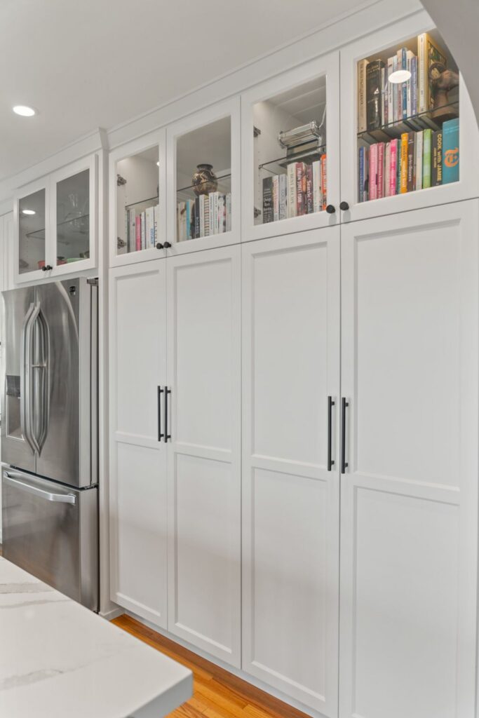 Floor-to-ceiling white pantry cabinetry with glass-front uppers, cookbook storage and stainless refrigerator.