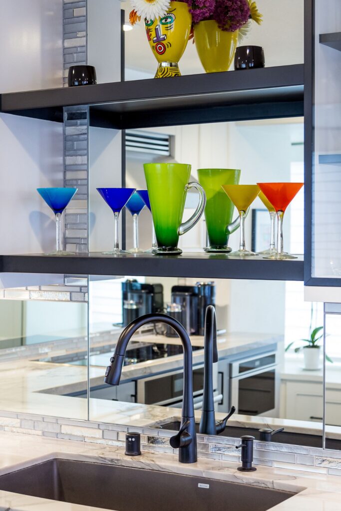 Kitchen sink with matte black faucet, mirrored backsplash and open shelves displaying colorful glassware.
