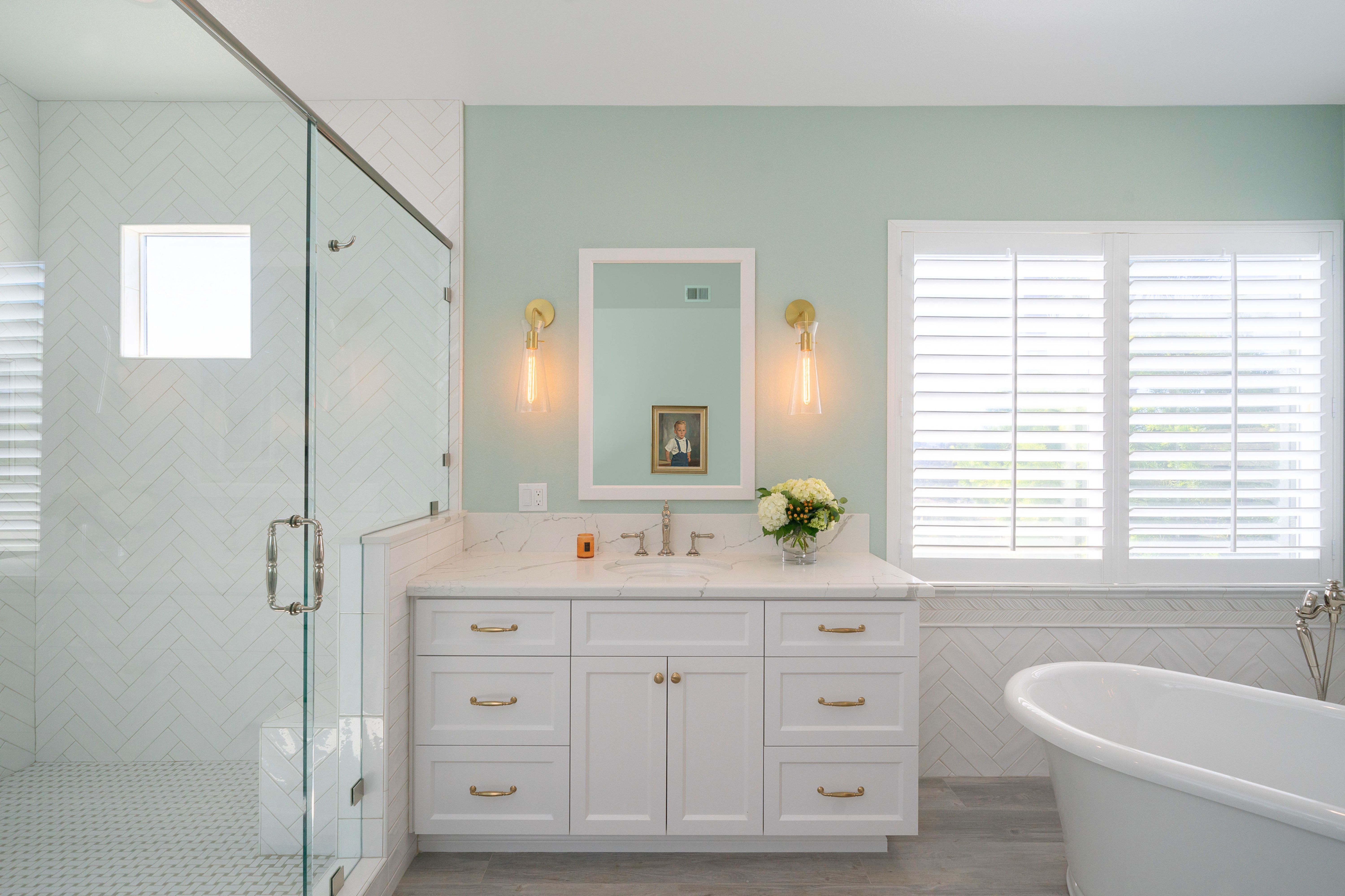 Primary bath with glass shower, white herringbone tile, freestanding tub, vanity and shuttered window.