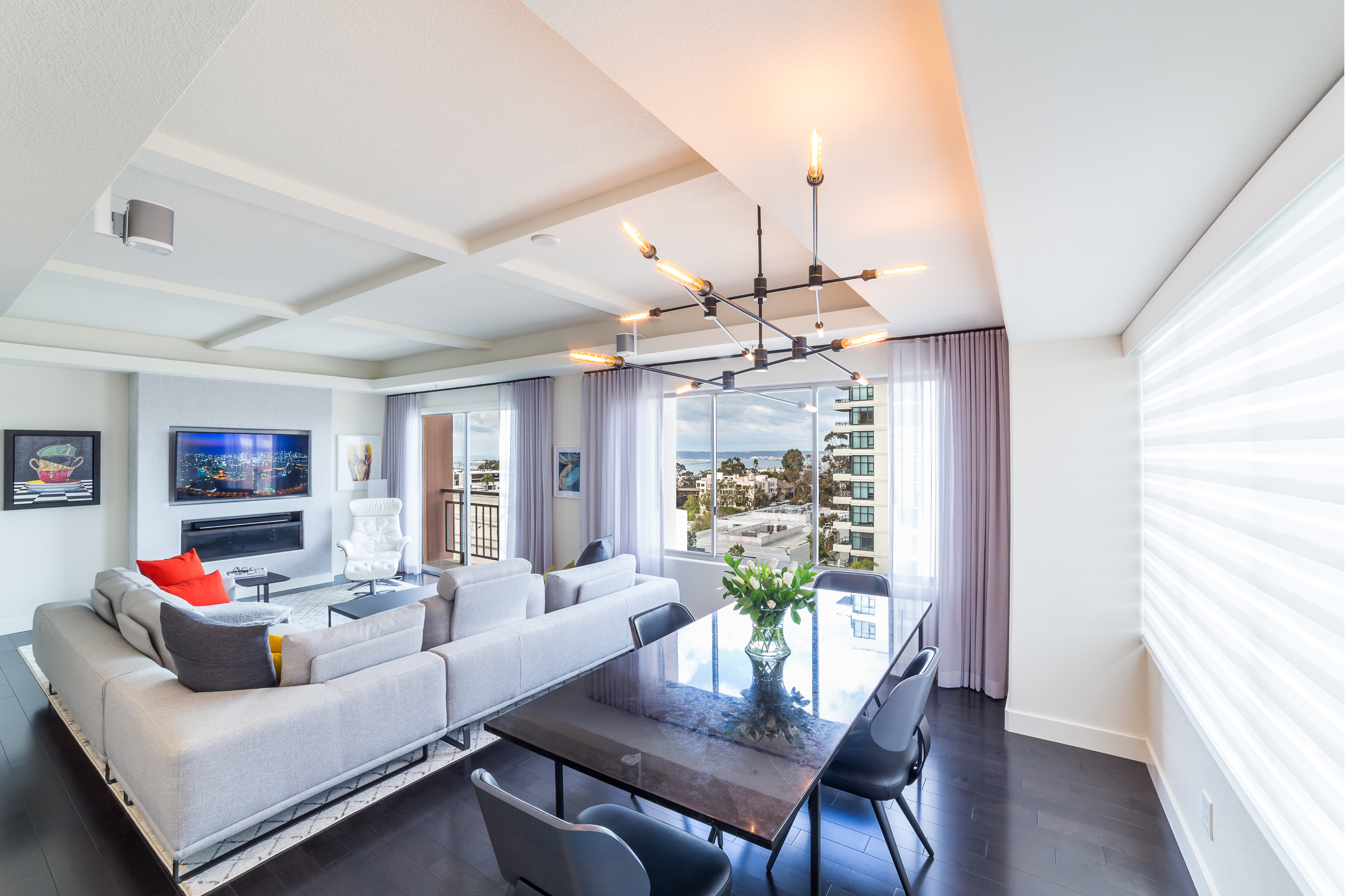Open living and dining area with dark wood floors, coffered ceiling, modern chandelier, fireplace and large windows.