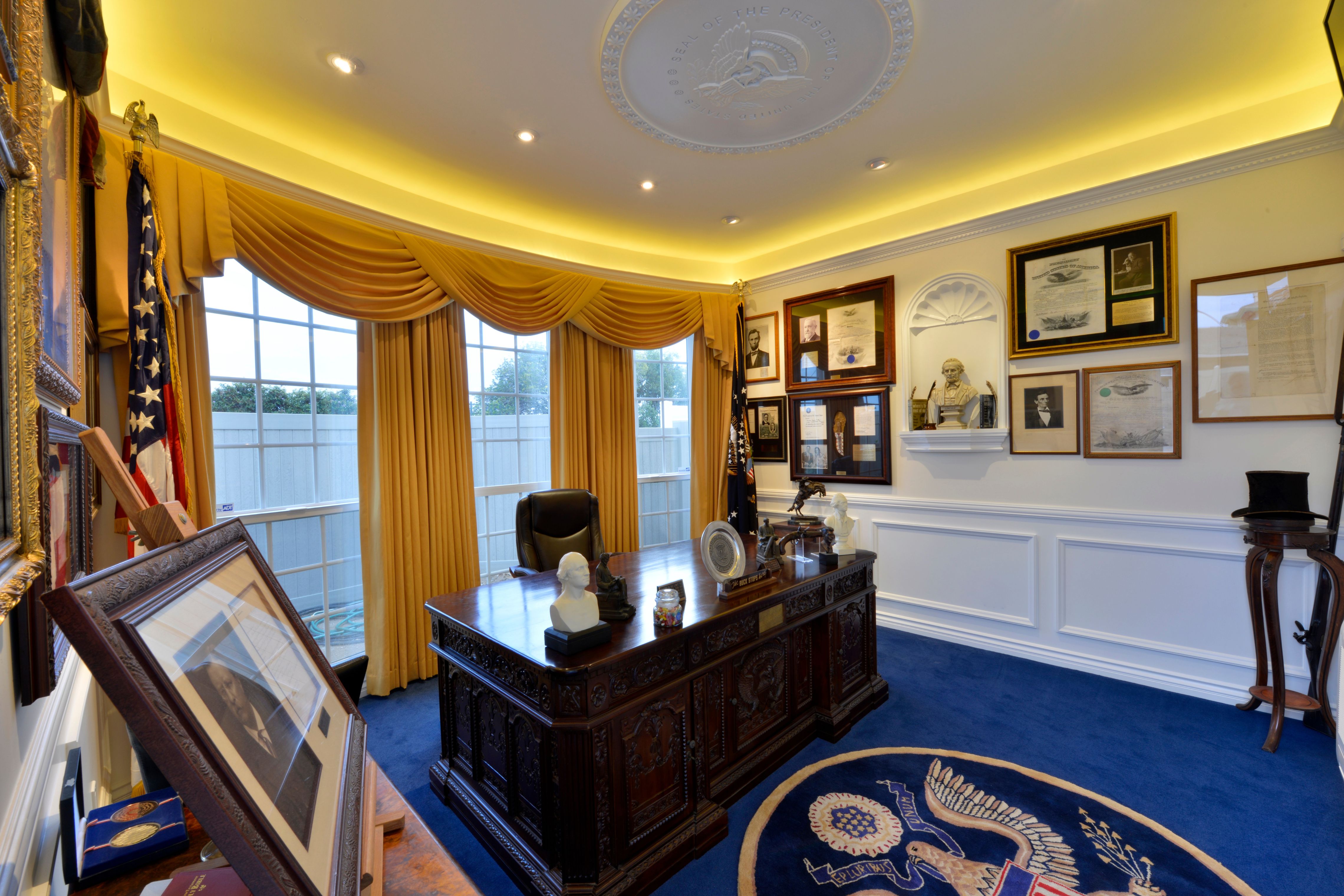 Home office with cove lighting, large windows, paneled walls, blue carpet and framed historical displays.