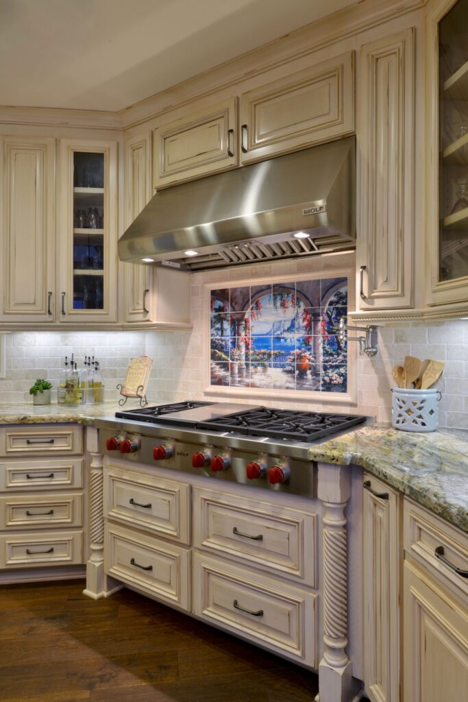 Wolf gas range with stainless hood, cream distressed cabinetry, granite counters and decorative tile mural.
